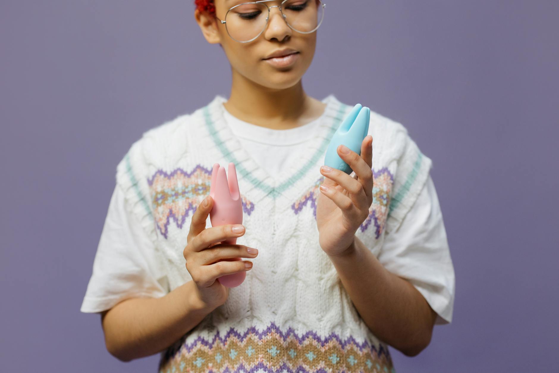 眼鏡をかけた女性が青とピンクのシリコンバイブレーターを思慮深く持っている