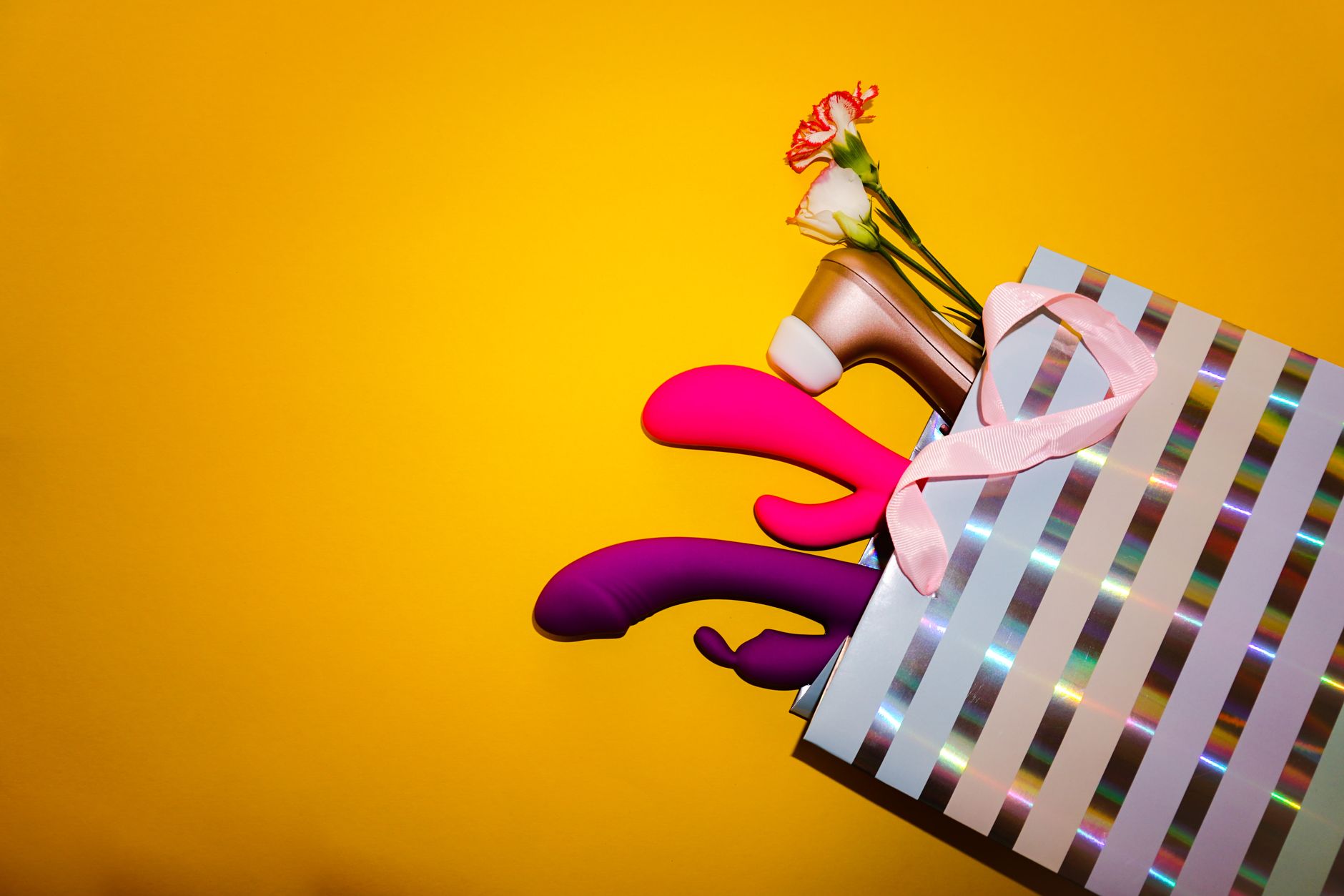Colorful vibrators with flowers in a holographic gift bag against a bold yellow background.